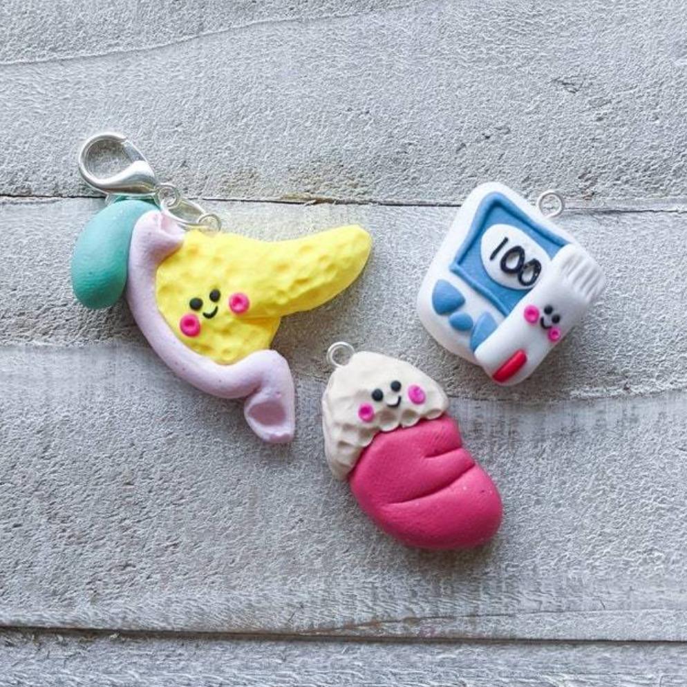 Three kawaii styled endocrinology charms – a yellow pancreas attached with duodenum and gallbladder, an adrenal gland on top of kidney, and glucose meter with a glucose strip.