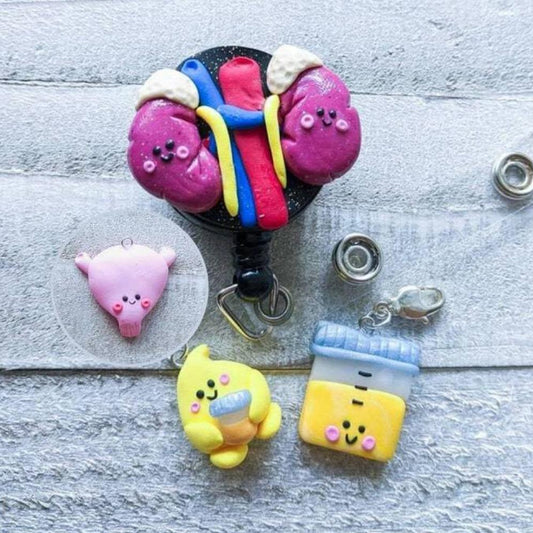 Renal charms are shown being used as a badge reel accessory. There are 3 kawaii styled charms - bladder, urine drop holding a urine analysis cup, and a filled urine analysis cup.